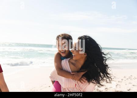 Happy biracial woman giving piggyback ride to daughter at beach against sky with copy space Stock Photo