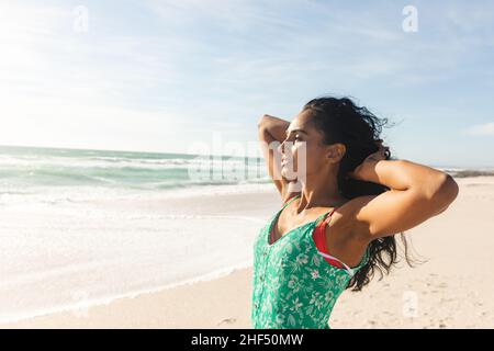 Young biracial woman with hands in hair looking away at beach against sky on sunny day Stock Photo