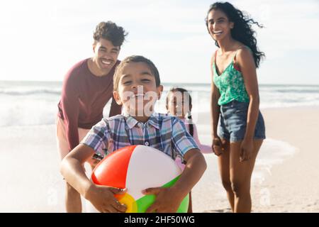 Portrait of smiling biracial boy holding ball with family at beach on sunny day Stock Photo