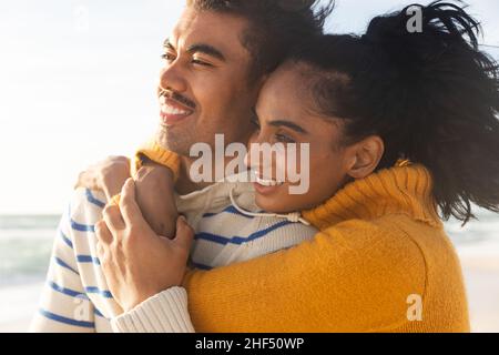Smiling biracial young woman embracing boyfriend from behind while looking away at beach Stock Photo