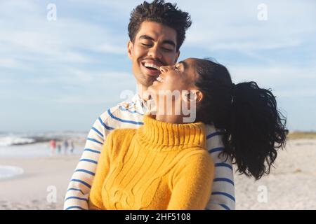Happy biracial man embracing cheerful young girlfriend from behind at beach against sky Stock Photo