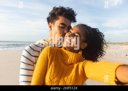 Cheerful young biracial woman taking selfie with man kissing her on cheek at beach Stock Photo