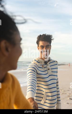 Smiling biracial man holding hand of girlfriend while standing behind her at beach against sky Stock Photo