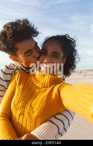 Portrait of smiling biracial woman taking selfie of boyfriend hugging and kissing on cheek at beach Stock Photo