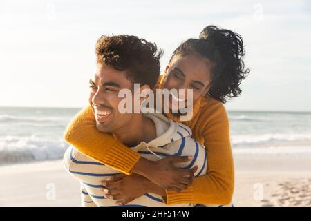 Cheerful biracial man giving piggyback ride to girlfriend at beach on sunny day Stock Photo