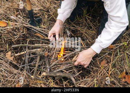 Close-up cropped shot of unrecognizable tourist man putting kindling on burning fire to keep warm and cook food at outdoors on overcast cold day. Stock Photo