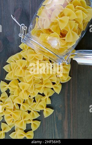Heap of Uncooked Farfalle Pasta Scattered from a Glass Jar on Black Wooden Table Stock Photo