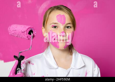 Young, cute girl standing in front of a pink colored wall. She is holding a roller filled with pink color and she has color in her face. Stock Photo