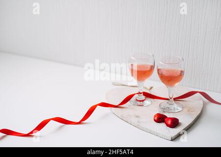 Valentines Day Festive table setting with two glasses of rose wine, red heart shape chocolate candies and red ribbons on white background. Valentine Stock Photo
