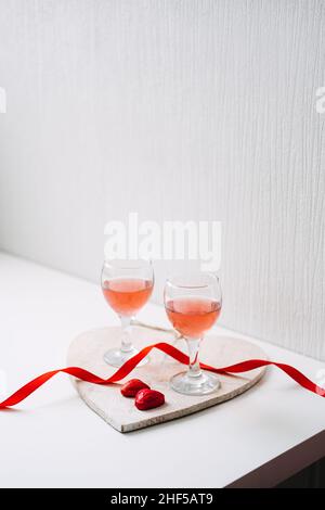 Valentines Day Festive table setting with two glasses of rose wine, red heart shape chocolate candies and red ribbons on white background. Valentine Stock Photo