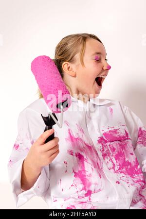 Young girl holding a roller filled with pink paint. She has color on her eyelid and is laughing happily. Stock Photo