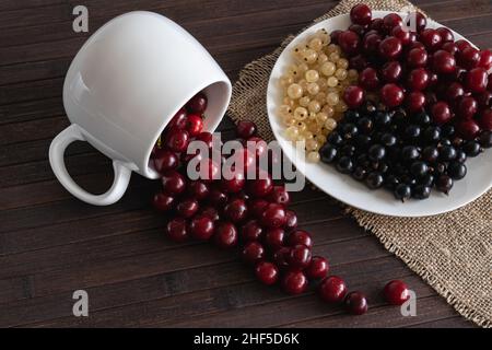 On a wooden table are cherries, white and black currants in a white porcelain mug and plate. Horizontal photo. Stock Photo
