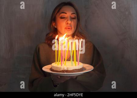 Young smiling woman holding homemade birthday cake with burning candles while standing indoors, side view. Female bringing Bday pie. Birthday traditio Stock Photo