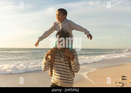 Happy biracial father carrying son on shoulders with arms outstretched enjoying sunset at beach Stock Photo