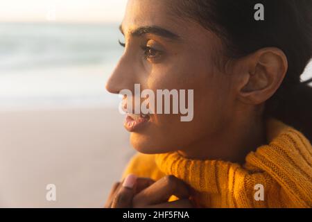 Close-up of young thoughtful biracial woman in sweater looking away at beach during sunset Stock Photo