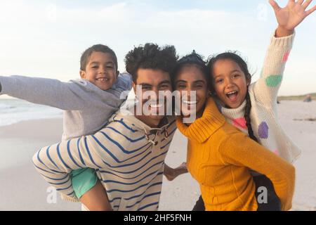 Portrait of happy biracial father and mother giving piggyback rides to kids at beach during sunset Stock Photo