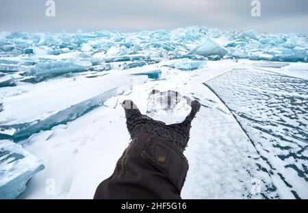 Open hand in glove holding small transparent piece of Ice at frozen lake with snow Stock Photo