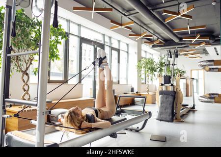 A woman is doing Pilates on a reformer bed in a bright studio. A