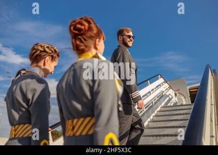 Smiling man pilot and two women flight attendants climbing plane boarding stairs under blue sky Stock Photo