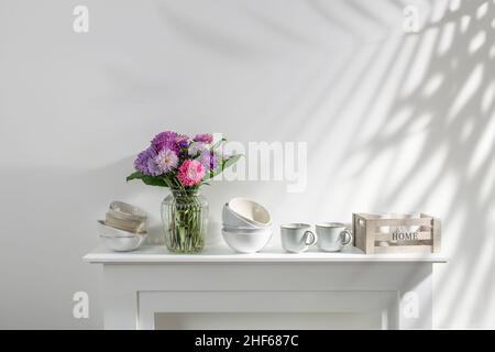 kitchen utensils, cups, bowls, bottles, storage items, a bouquet of asters are on a white dresser Stock Photo