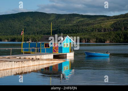 A small boathouse at the end of a wooden pier. The bright teal blue building has a red door and yellow trim. There's a small rowboat anchored to pier. Stock Photo