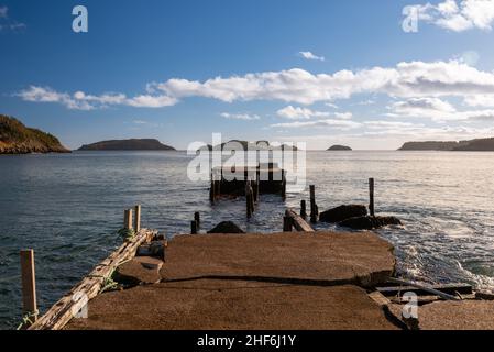 Old wooden weather beaten and worn wharf at the edge of the ocean on a bright blue sunny day with white clouds. The concrete deck is broken in pieces. Stock Photo