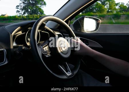 Durham, UK - 23rd August 2019: Confident male driver holding the steering wheel of a Mazda vehicle. Sunshine and trees reflect in the side mirror Stock Photo