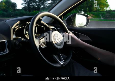 Durham, UK - 23rd August 2019: Confident male driver holding the steering wheel of a Mazda vehicle. Sunshine and trees reflect in the side mirror Stock Photo