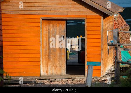 Orange wooden fishing shed for storing fishing gear and boats. The building has a worn wooden door, window, peaked roof, fishing rope, and gear. Stock Photo