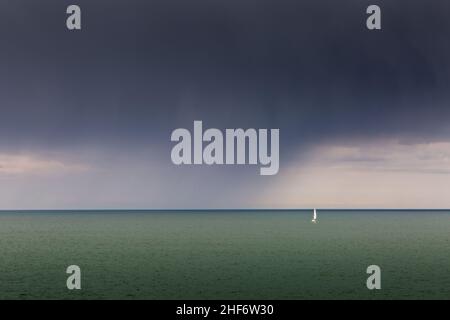 a small sailing boat with a white sail is all alone in the sea, as rain clouds draw nearer Stock Photo