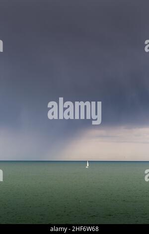 a small sailing boat with a white sail is all alone in the sea, as rain clouds draw nearer Stock Photo