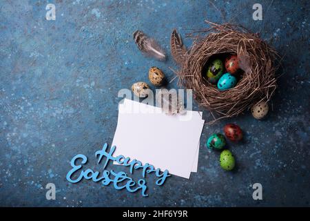 Small colorful painted quail eggs as symbol of Easter day in bird nest as traditional decoration and mock-up of card on blue concrete background Stock Photo