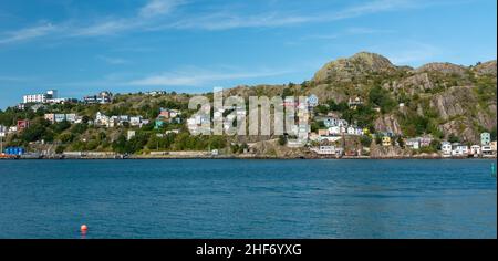 Aerial view of St. John's Harbour, Newfoundland, on a sunny day, under blue sky and white clouds. The colorful wooden houses are scattered on a hill. Stock Photo