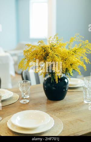 Wooden table with empty plates and glasses. Festive Easter table setting. Branches of mimosa in vase. Stock Photo