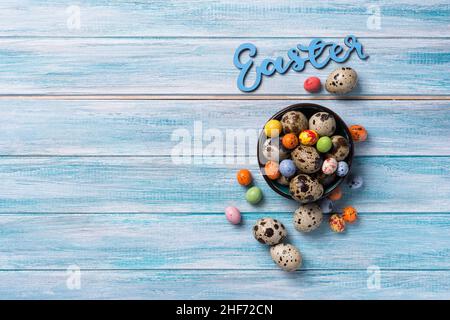 Top view of small quail eggs and colored candies in bowl for Easter on blue wooden background with copy space Stock Photo