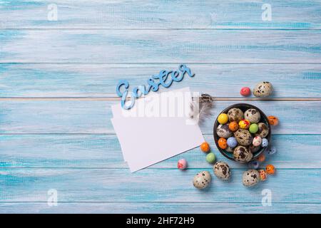 Top view of small quail eggs, colored candies and mock-up of blank white card for Easter on blue wooden background Stock Photo
