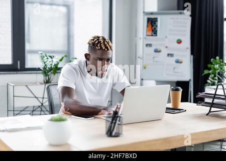 young african american man with vitiligo working on laptop near paper cup in office Stock Photo
