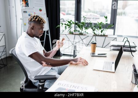 young african american man with vitiligo using cellphone near laptop and paper cup in office Stock Photo
