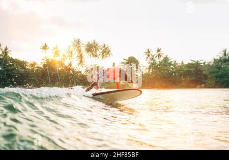 Black long-haired teen boy riding and jumping from a long surfboard. He caught a  wave in an Indian ocean bay with magic sunset background. Extreme wa Stock Photo