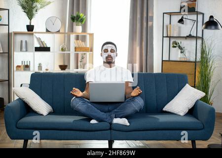Front view of handsome peaceful African American bearded man with white facial mask, sitting in lotus position on sofa with laptop, meditating with eyes closed Stock Photo