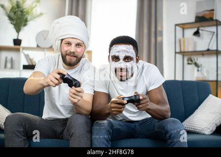 Multiethnic gay couple at home. Two emotional smiling multiracial male friends with facial masks and eye patches sitting on the couch and playing video games using joysticks Stock Photo