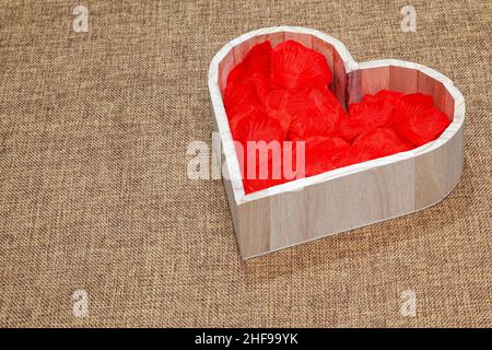 A heart-shaped wooden tray with red petals made of fabric inside, placed on a sackcloth surface. Stock Photo