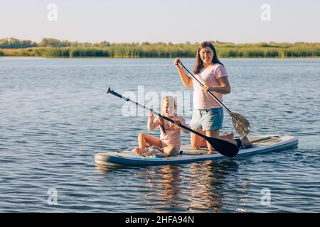 Happy Caucasian mother and little daughter sup boarding with oars in hands looking at camera and smiling on crystal blue lake with green reeds in Stock Photo