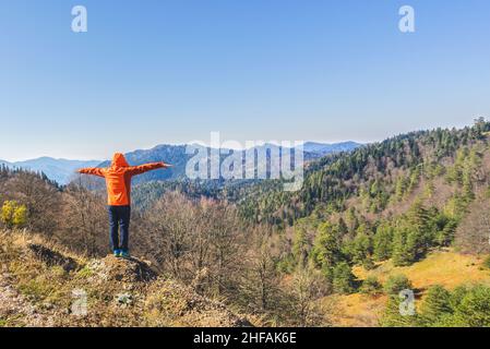 Man is wearing orange hoodie, standing on the hill with open arms and looking at forest. Autumn view of Yedigoller National Park in Turkey. Stock Photo