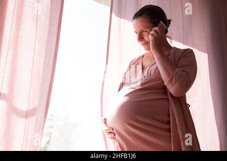 Young pregnant woman calling on the phone standing near window Stock Photo