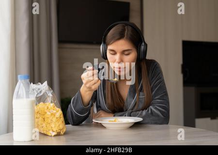 Young woman in pajamas having breakfast with corn flakes with milk at home. Listen to music with headphones. copy space Stock Photo