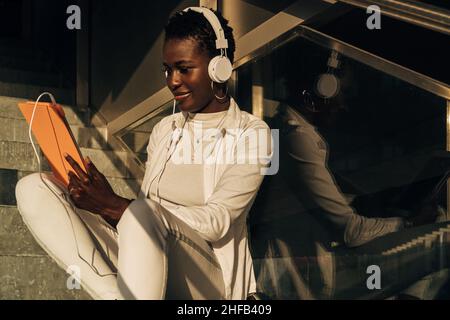 Black woman using digital tablet with headphones while sitting relaxed on stairs. Stock Photo