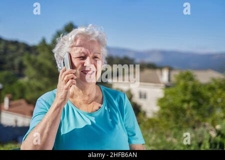Positive elderly female with gray hair having conversation on mobile phone in sunny day against green mountains with cottages Stock Photo