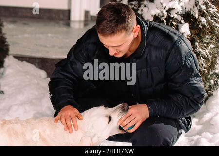 A man in winter clothes outside taking a blue little ball from the dog mouth to continue playing with the dog. Stock Photo