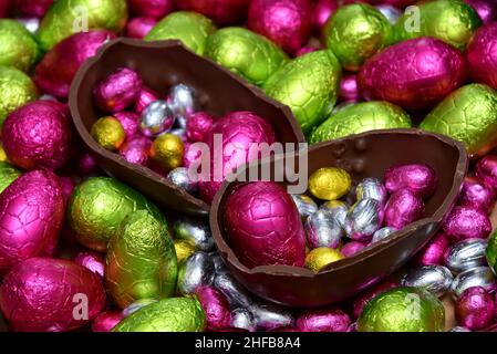Pile of multi colored and different sizes of colourful foil wrapped chocolate easter eggs in pink, red, gold, yellow and lime green with two halves of Stock Photo